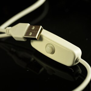 May include: Close-up of a white USB cable with a USB connector and an inline on/off switch. The cable and switch are against a black background, with the cable slightly out of focus.