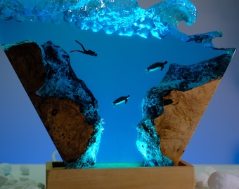 Könnte beinhalten: Eine Harzskulptur einer Welle in Blau- und Grüntönen. Die Welle ist hohl und enthält eine Szene mit einem Taucher und Meeresschildkröten, die in einem blauen Harz schwimmen. Die Skulptur wird von unten mit einem blauen Licht beleuchtet. Die Skulptur steht auf einem Holzsockel.