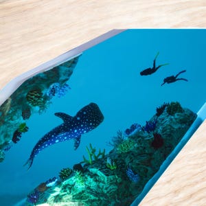 May include: A resin art piece depicting a whale shark swimming in a blue ocean with coral reefs and two scuba divers in the background.
