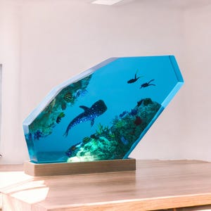 May include: A blue resin sculpture of a whale shark swimming in a coral reef. The sculpture is on a wooden base.