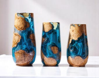 Puede incluir: Tres jarrones decorativos con un diseño único. Presentan una combinación de madera marrón y resina azul, creando un contraste llamativo. La resina parece fluir como agua, añadiendo un elemento dinámico. Los jarrones son de diferentes alturas.
