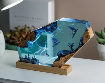 Puede incluir: Una escultura de resina azul con una base de madera. La resina presenta una escena de un castillo y dragones volando. La escultura está iluminada desde atrás, creando un efecto brillante.