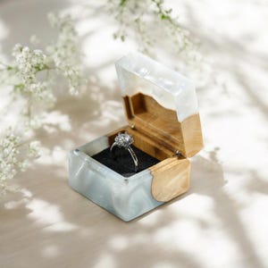 Puede incluir: Una caja de anillo de madera y blanca con un anillo dentro. El anillo tiene una piedra grande y transparente en el centro.