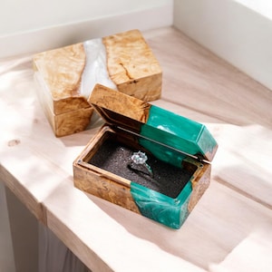 Puede incluir: Una caja de madera para anillos con una tapa de resina verde. La caja está abierta y hay un anillo de diamantes dentro.