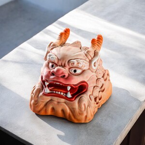 Pu&ograve; includere: Una scultura in ceramica di un leone guardiano mitico giapponese, noto come Komainu, con un'espressione feroce e dettagli dipinti in rosso. La scultura &egrave; di colore marrone chiaro e ha una superficie testurizzata.