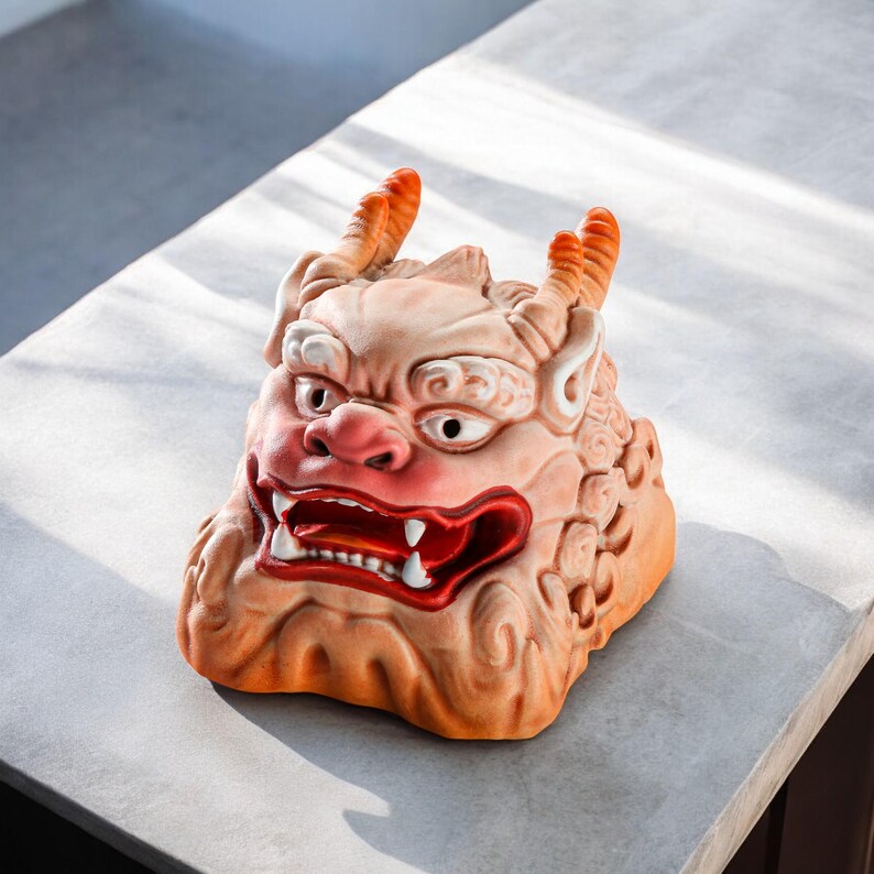 Pu&ograve; includere: Una scultura in ceramica di un leone guardiano mitico giapponese, noto come Komainu, con un'espressione feroce e dettagli dipinti in rosso. La scultura &egrave; di colore marrone chiaro e ha una superficie testurizzata.