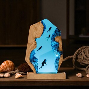 May include: A decorative wooden lamp featuring a blue resin center with sharks and divers. The lamp has a wooden base and a natural wood grain border. The light shines through the resin, creating a unique underwater scene. The lamp is a decorative item.