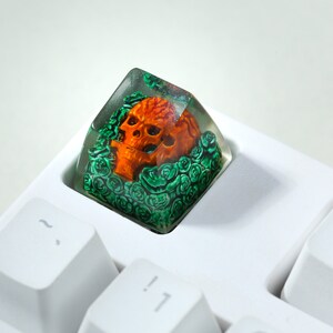 May include: A clear resin keycap with a detailed orange skull surrounded by green foliage. The keycap is sitting on a white keyboard.