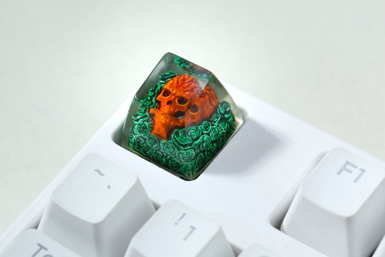 May include: A clear resin keycap with a detailed orange skull surrounded by green foliage. The keycap is sitting on a white keyboard.
