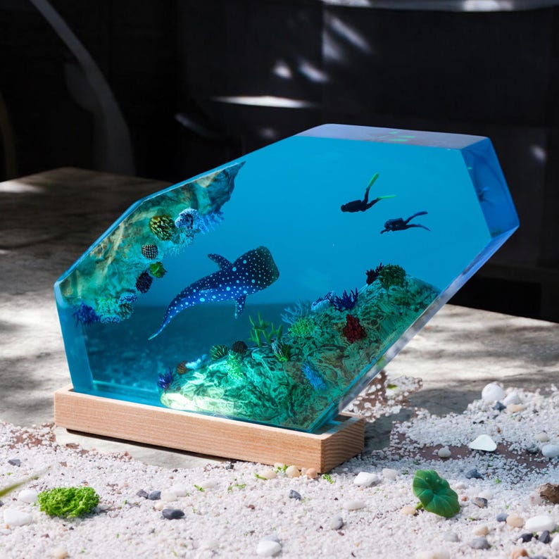 May include: A clear blue resin sculpture of an underwater scene with a whale shark, coral, and small fish. The sculpture is on a wooden base.