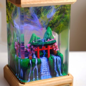 Könnte beinhalten: Eine Miniatur-Szene eines japanischen Gartens, die in einem durchsichtigen Harz-Würfel eingeschlossen ist. Die Szene zeigt ein rotes Torii-Tor, einen Wasserfall, grüne Hügel und rosa Kirschblüten. Der Würfel ist auf einem Holzsockel montiert.