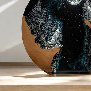 Puede incluir: Una obra de arte redonda de madera que representa una escena nocturna con un lobo aullando. La obra presenta un fondo negro con estrellas blancas, una luna llena y pinos blancos. El lobo es blanco y la madera tiene un acabado marr&oacute;n natural.