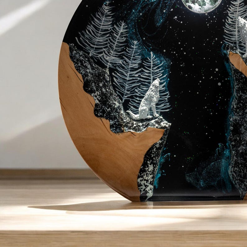 Puede incluir: Una obra de arte redonda de madera que representa una escena nocturna con un lobo aullando. La obra presenta un fondo negro con estrellas blancas, una luna llena y pinos blancos. El lobo es blanco y la madera tiene un acabado marr&oacute;n natural.