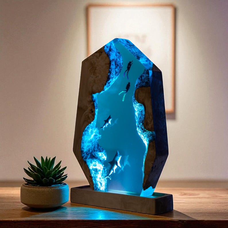 May include: A decorative resin lamp featuring a blue underwater scene with divers and sharks. The lamp has a geometric shape with a wooden exterior and a glowing blue interior. The lamp sits on a rectangular base.