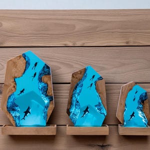 May include: Three wooden sculptures with a blue resin center depicting an underwater scene with sharks and divers. The sculptures are set on small wooden shelves against a wood-paneled wall. The resin is a vibrant blue.