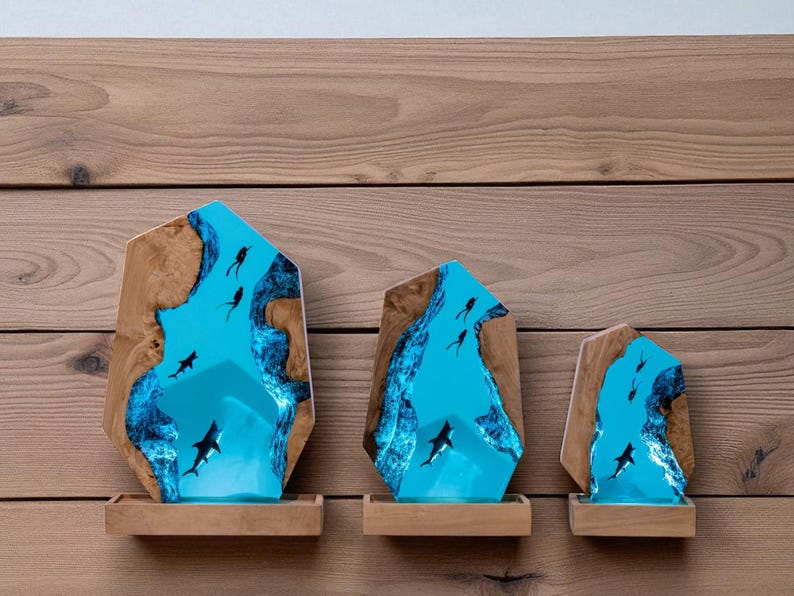 May include: Three wooden sculptures with a blue resin center depicting an underwater scene with sharks and divers. The sculptures are set on small wooden shelves against a wood-paneled wall. The resin is a vibrant blue.