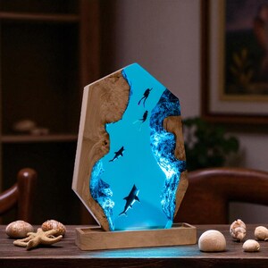 May include: A decorative wooden lamp with a blue resin center depicting an underwater scene with sharks and divers. The lamp has a rectangular wooden base and a unique, irregular shape. The blue resin glows, creating a captivating visual effect.