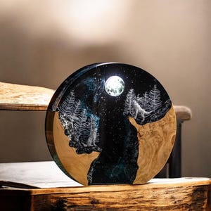 Puede incluir: Una obra de arte redonda de madera que representa una escena nocturna con un lobo aullando a la luna. La pieza tiene un fondo de resina azul oscuro y negro con &aacute;rboles blancos y una luna llena. La base es de madera clara.