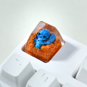 May include: A blue skull keycap with orange and brown resin details, encased in a clear resin cube. The keycap is sitting on a white keyboard.