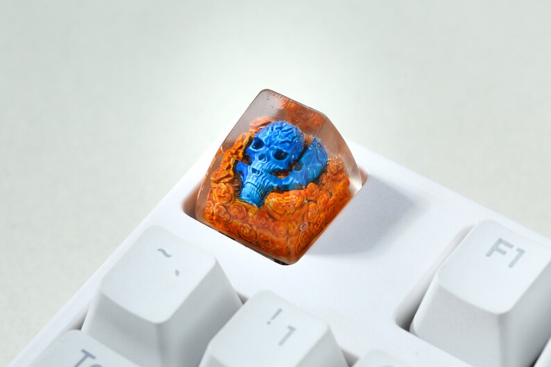 May include: A blue skull keycap with orange and brown resin details, encased in a clear resin cube. The keycap is sitting on a white keyboard.