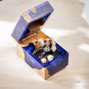 Puede incluir: Una caja de anillo de madera y azul con un anillo y flores secas dentro. La caja está abierta y el anillo es visible.