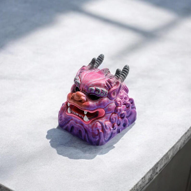 Pu&ograve; includere: Un copritasto in ceramica viola e rosa a forma di creatura mitologica con corna e un'espressione feroce. Il copritasto presenta dettagli intricati e una superficie testurizzata.