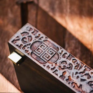 May include: A wooden box with a carved design featuring a geometric pattern and intricate details. The box has a brass hinge and a dark brown finish.