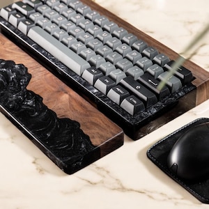 以下が含まれることがあります： 黒と灰色のメカニカルキーボードと、クルミ材と黒の樹脂製のリストレスト。黒いマウスは黒い革製のマウスパッドの上にある。