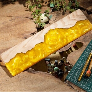 May include: A wooden wrist rest with a yellow resin inlay. The resin has a swirling, wave-like pattern. The wrist rest is on a wooden surface.