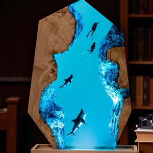 May include: A decorative wooden art piece featuring a vibrant blue resin river scene with sharks and scuba divers. The wooden frame has a natural wood grain. The resin river glows with a blue light, creating a unique underwater effect.