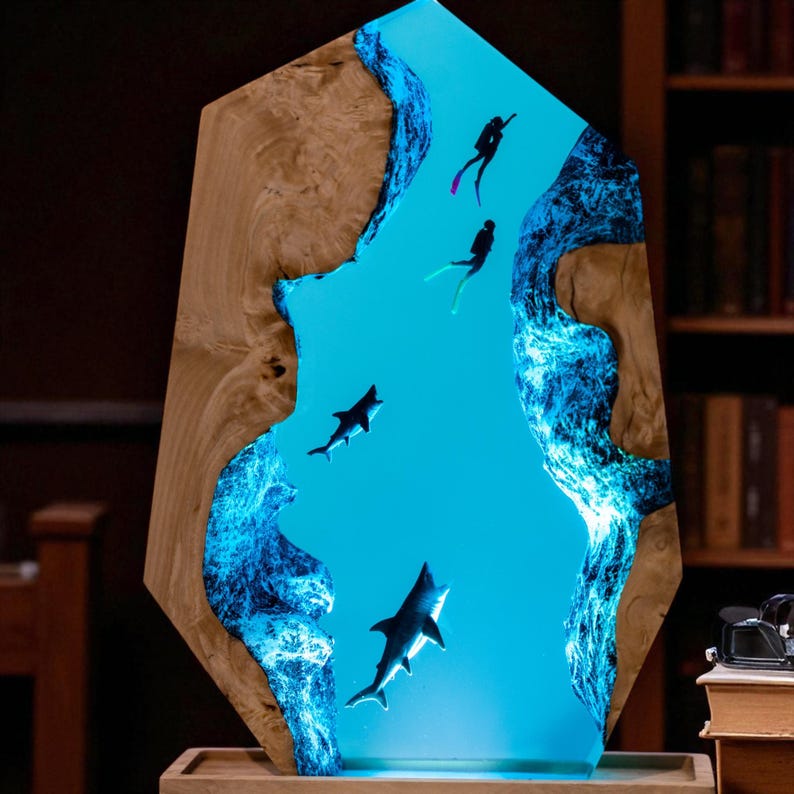 May include: A decorative wooden art piece featuring a vibrant blue resin river scene with sharks and scuba divers. The wooden frame has a natural wood grain. The resin river glows with a blue light, creating a unique underwater effect.