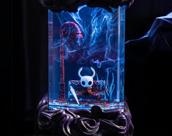 Puede incluir: Una cúpula de vidrio con una base negra, que contiene una figura en miniatura del Caballero del videojuego Hollow Knight. El Caballero está sentado en un banco con una espada a su lado. La cúpula está llena de humo azul.