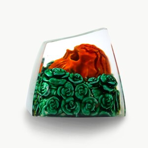 May include: A clear glass cube with a sculpted orange skull surrounded by green roses.