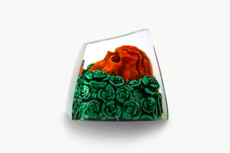 May include: A clear glass cube with a sculpted orange skull surrounded by green roses.