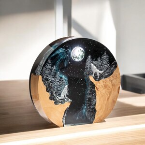 Puede incluir: Objeto decorativo redondo con una escena nocturna. El dise&ntilde;o presenta una luna llena, estrellas y lobos blancos aullando a la luna. La base es de madera clara y el cielo es azul oscuro con detalles blancos.
