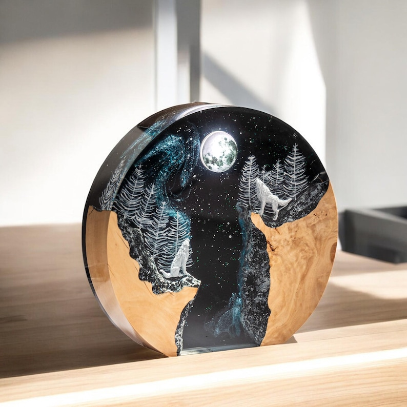 Puede incluir: Objeto decorativo redondo con una escena nocturna. El dise&ntilde;o presenta una luna llena, estrellas y lobos blancos aullando a la luna. La base es de madera clara y el cielo es azul oscuro con detalles blancos.