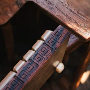 May include: Close-up of a wooden chair with a decorative geometric pattern carved into the armrest. The chair is made of dark wood with a natural finish.