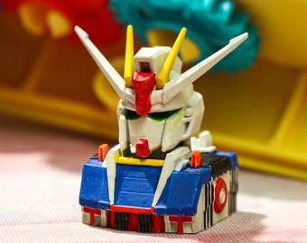May include: A small, white, blue, and red robot toy with a yellow antenna. The robot has a detailed face with green eyes and a red mouth. The robot is sitting on a blue and white base.