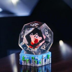 Può includere: Un dado trasparente a più facce con un personaggio rosa e rosso al suo interno. Il dado è appoggiato su una base che sembra uno skyline urbano.
