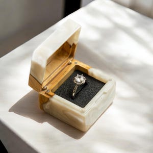 Puede incluir: Una caja de anillo blanca y marrón con un anillo de diamantes en su interior. El anillo está engastado en una banda de plata con un halo de diamantes más pequeños.