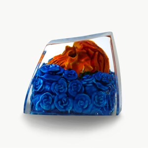 May include: A clear glass cube with a small orange skull resting on a bed of blue roses.
