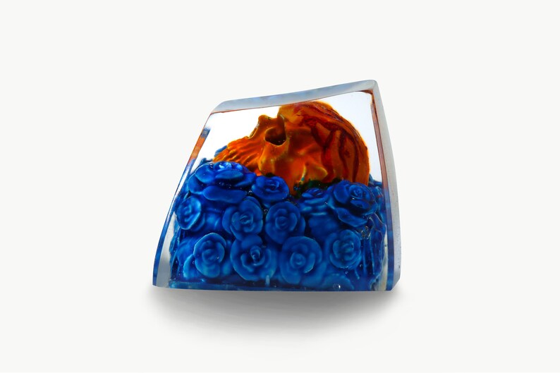 May include: A clear glass cube with a small orange skull resting on a bed of blue roses.