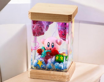 May include: A pink and white resin figurine of a video game character, Kirby, inside a clear glass cube with a wooden base. The cube also contains colorful resin swirls, stars, and other small figurines.