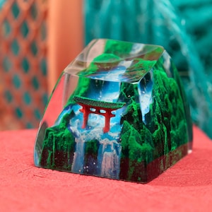 May include: A clear resin keycap with a miniature red torii gate and a green waterfall scene inside. The keycap is on a red surface.