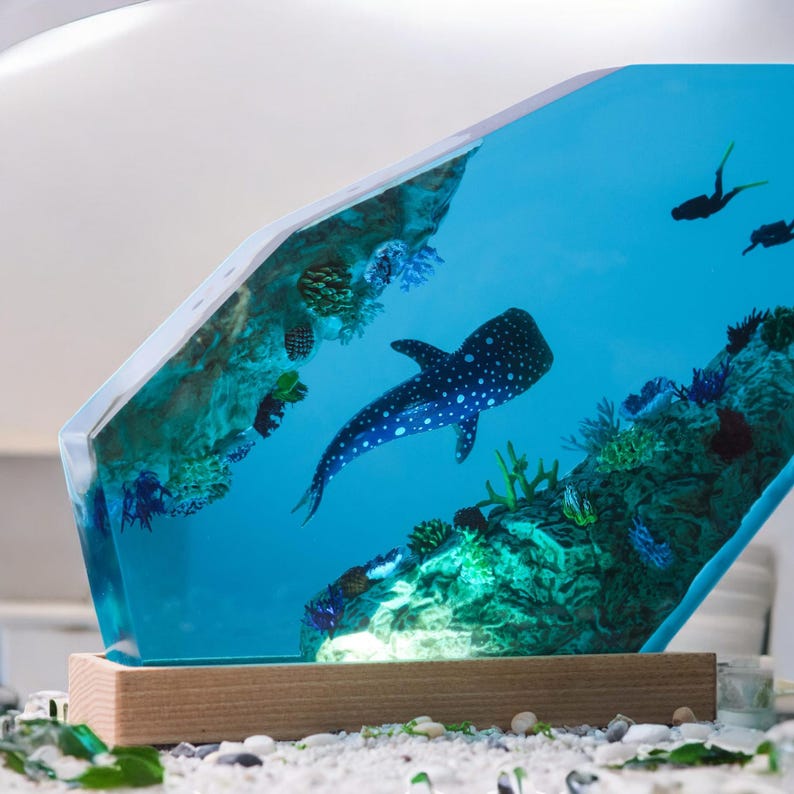 May include: A blue resin sculpture of a whale shark swimming in a coral reef. The sculpture is on a wooden base and has a light inside.
