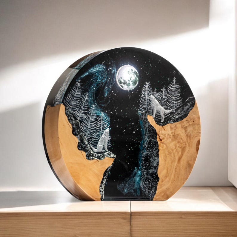 Puede incluir: Escultura de madera redonda que representa una escena nocturna con luna llena, estrellas y un lobo aullando. La obra de arte combina la veta natural de la madera con una resina oscura y brillante, creando un contraste llamativo. La pieza se exhibe sobre una superficie de madera.