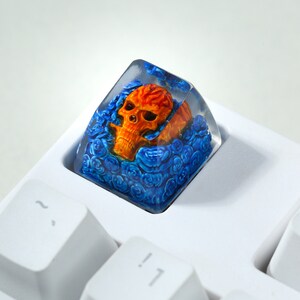 May include: A clear resin keycap with a skull design. The skull is orange and yellow with blue roses surrounding it.