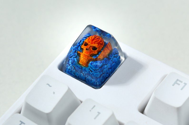 May include: A clear resin keycap with a skull design. The skull is orange and yellow with blue roses surrounding it.
