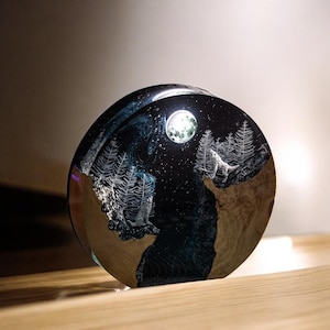 Puede incluir: Una pieza de arte de resina redonda que representa una escena nocturna con un lobo aullando a la luna. La obra de arte incluye &aacute;rboles grabados, un cielo estrellado y un r&iacute;o azul oscuro. La base tiene un aspecto de vetas de madera.
