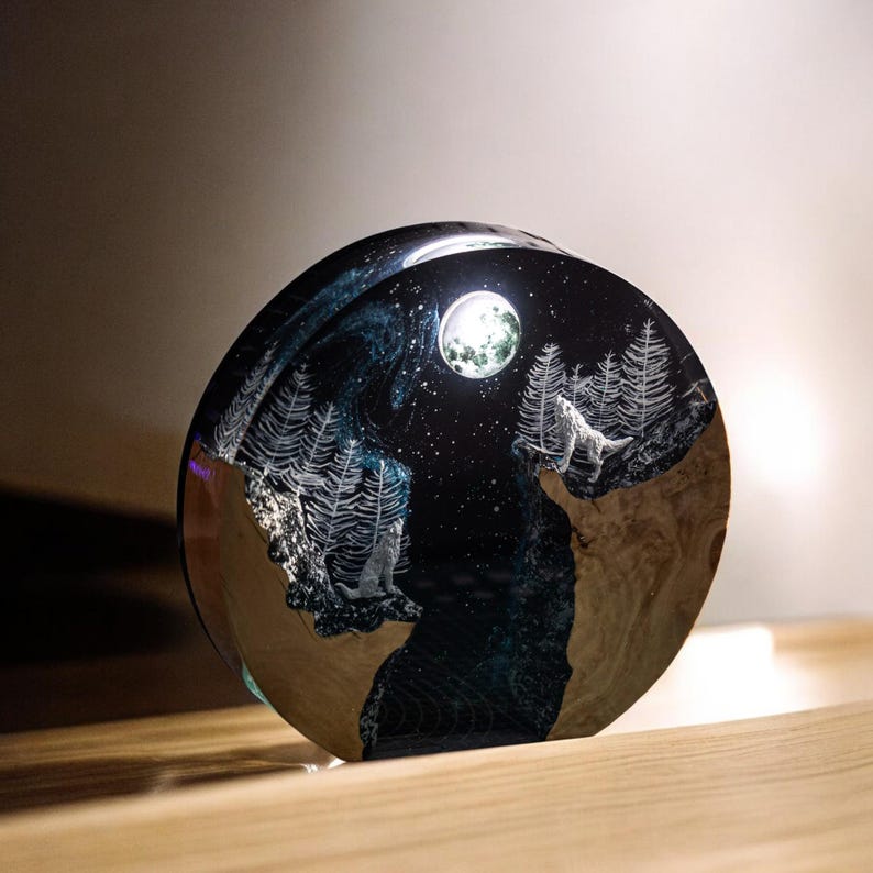 Puede incluir: Una pieza de arte de resina redonda que representa una escena nocturna con un lobo aullando a la luna. La obra de arte incluye &aacute;rboles grabados, un cielo estrellado y un r&iacute;o azul oscuro. La base tiene un aspecto de vetas de madera.